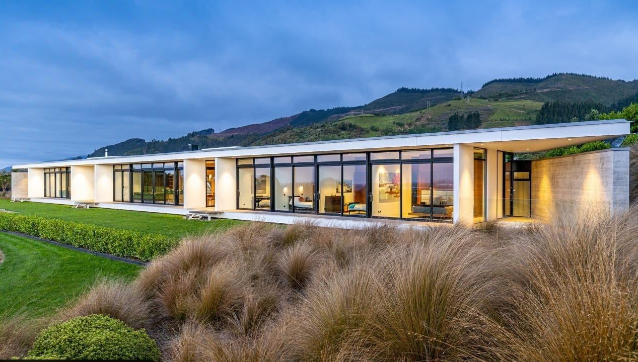 Single level home at Common Ground lit at dusk with tussock planting and North Canterbury hills behind